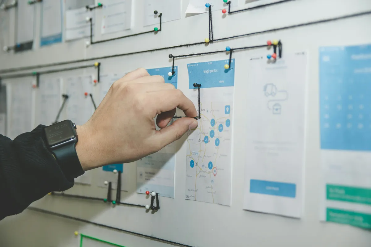 Hand pinning yarn between printed app screens on a wall-mounted flow diagram