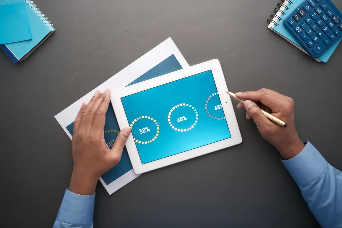 Overhead view of hands holding a tablet that shows percentage progress rings on a dark desk