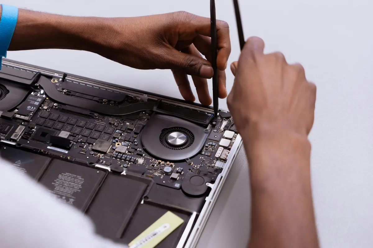 Hands using pry tools on an open laptop with its logic board, cooling fans and battery exposed